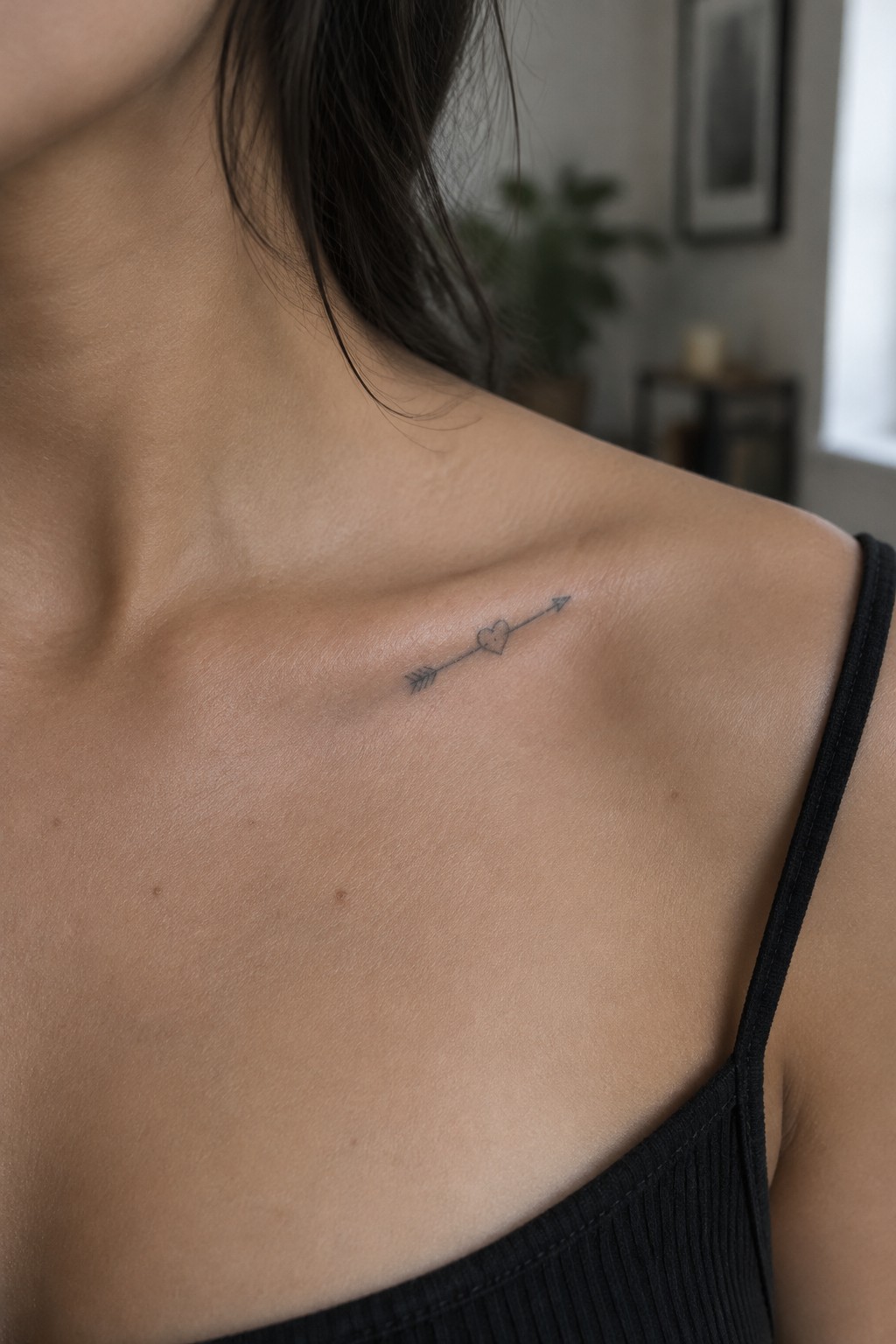 Small fine-line black heart and arrow tattoo on a woman’s collarbone.