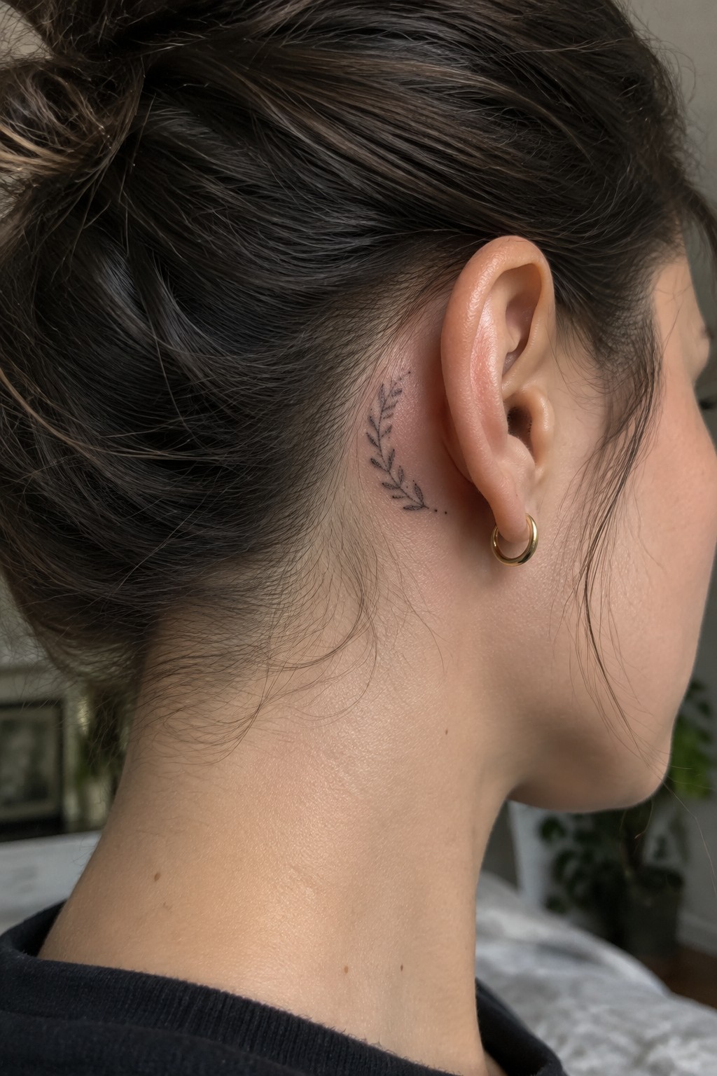Small black leafy sprig tattoo with dotted accents behind a woman’s ear.