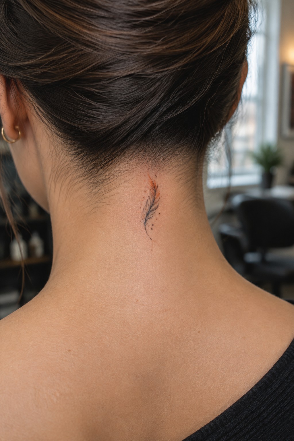 Small fine-line feather tattoo with dot details centered on the nape of a woman’s neck.
