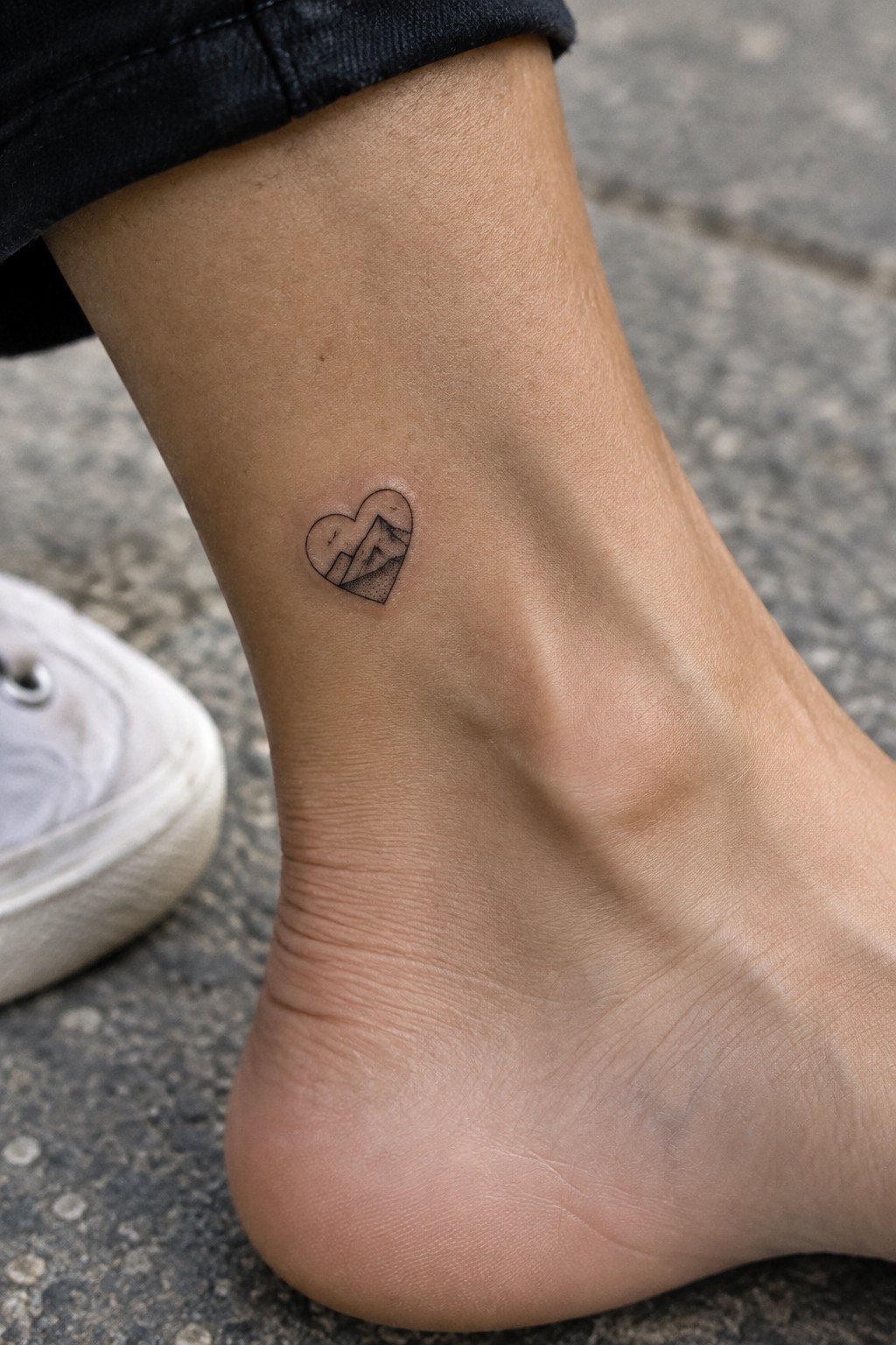 Small black linework tattoo of a mountain landscape inside a heart on the side of an ankle.