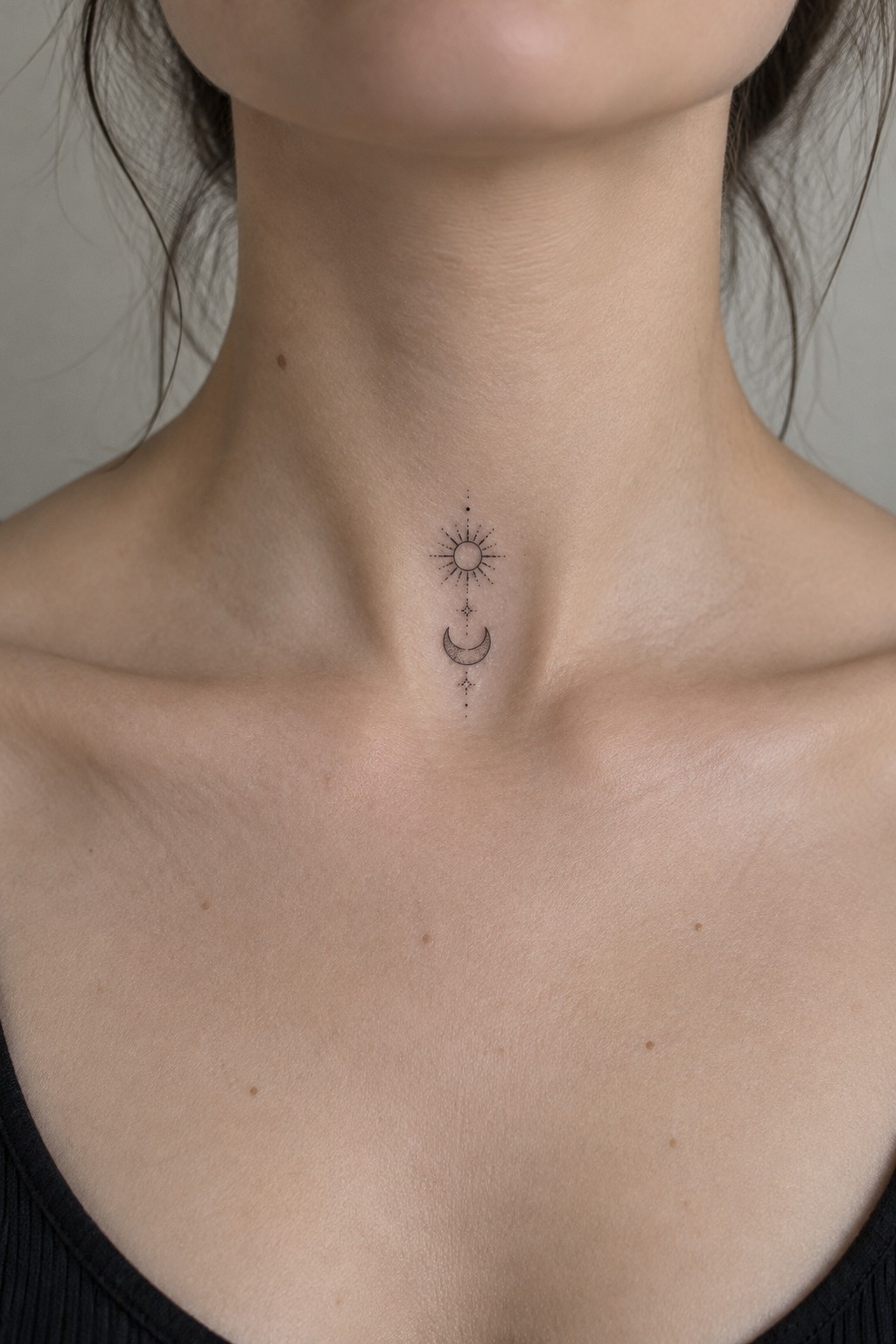 Small black sun and crescent moon tattoo aligned vertically at the center of a woman’s upper chest.