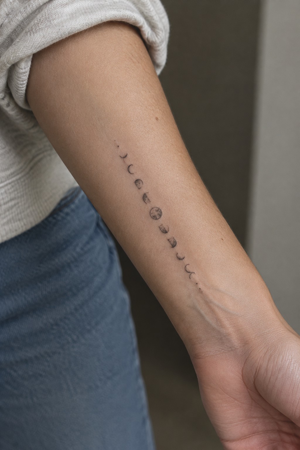 Fine line tattoo of the moon phases running lengthwise down the inner forearm.