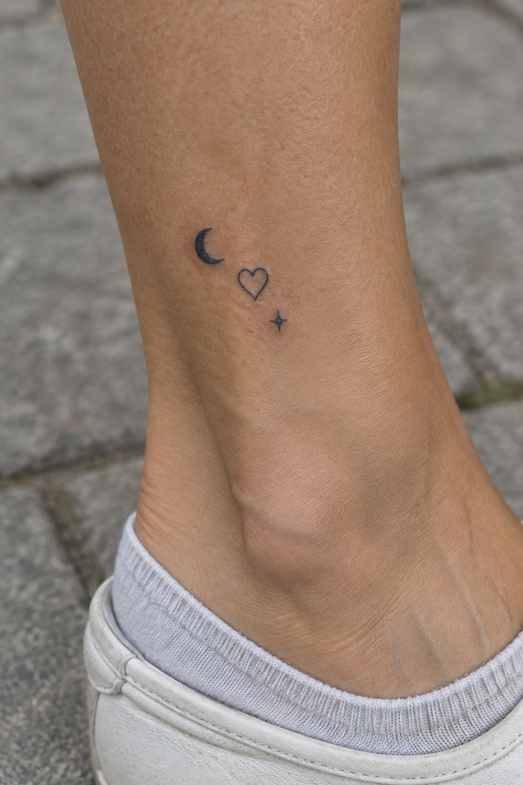 Small black crescent moon, outlined heart, and star tattoo on the inner ankle above a white sneaker.