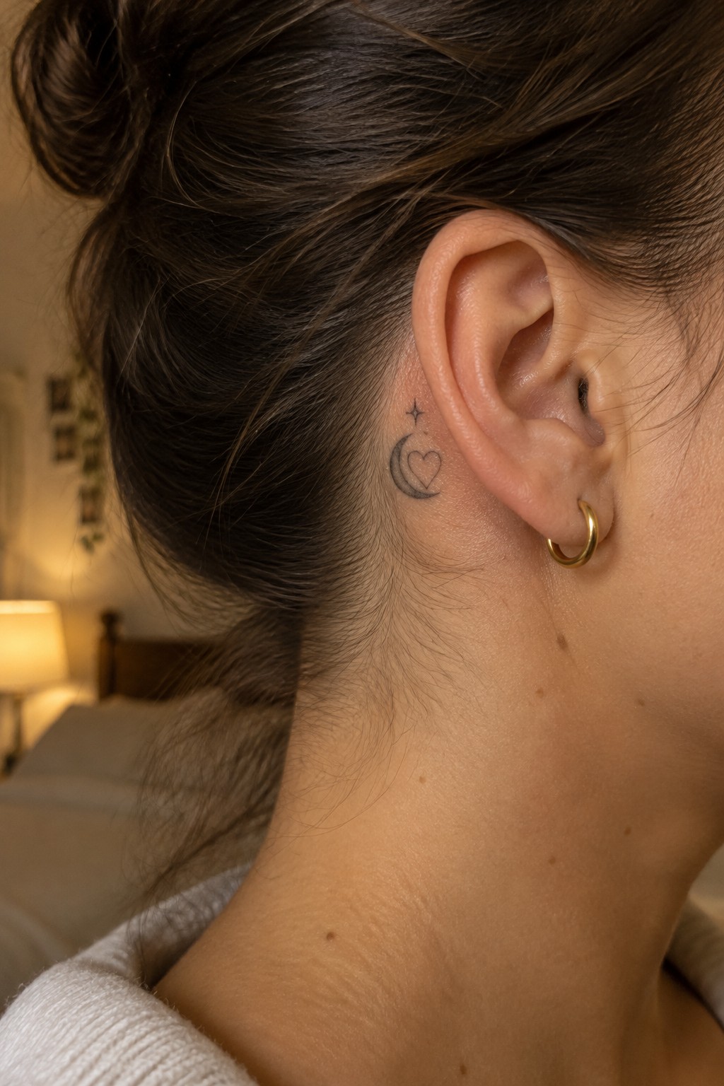 Small fine-line tattoo of a crescent moon, outlined heart, and star behind a woman’s ear.