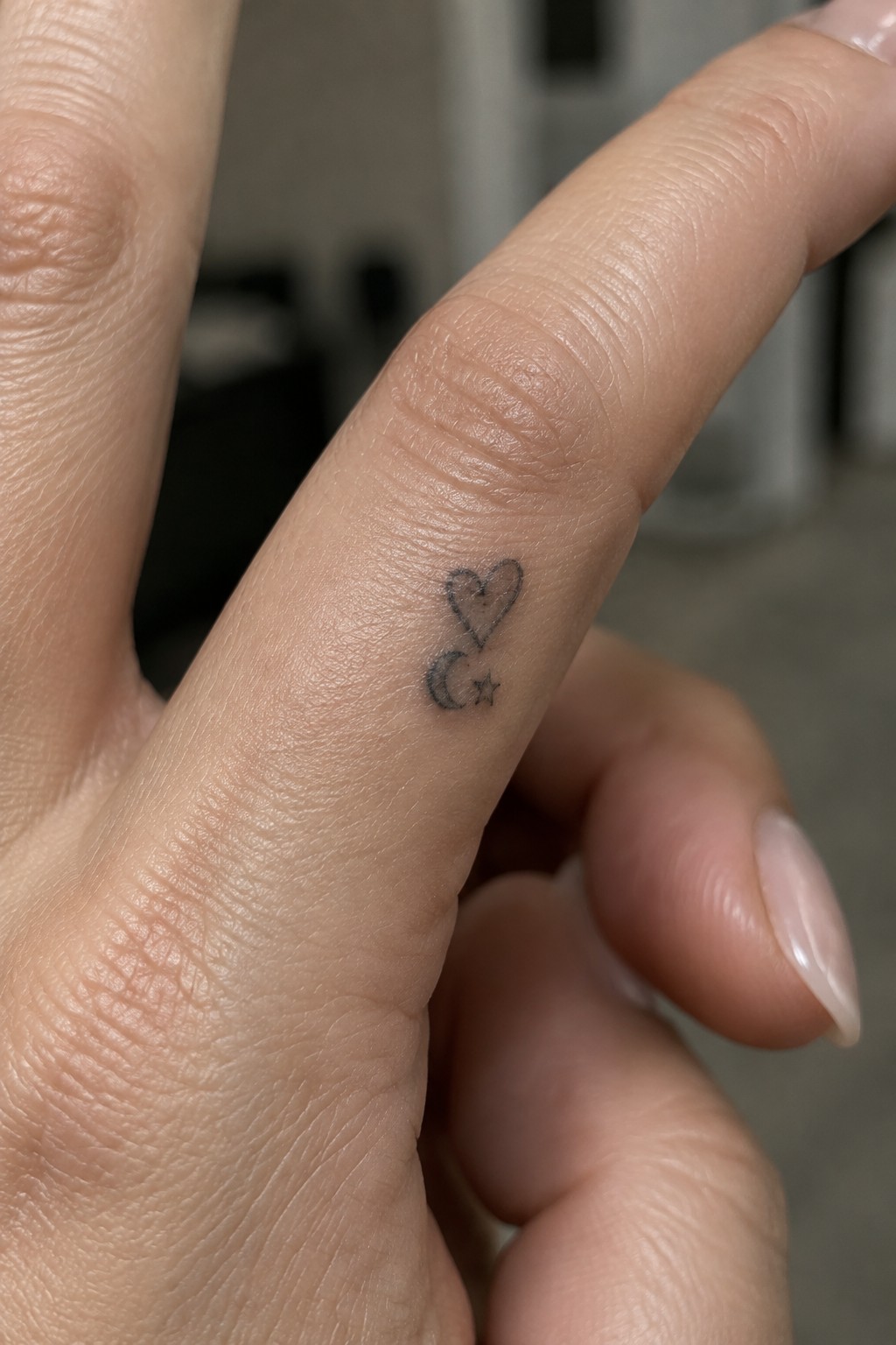 Small black outline tattoo of a heart, crescent moon, and star on the side of a finger.