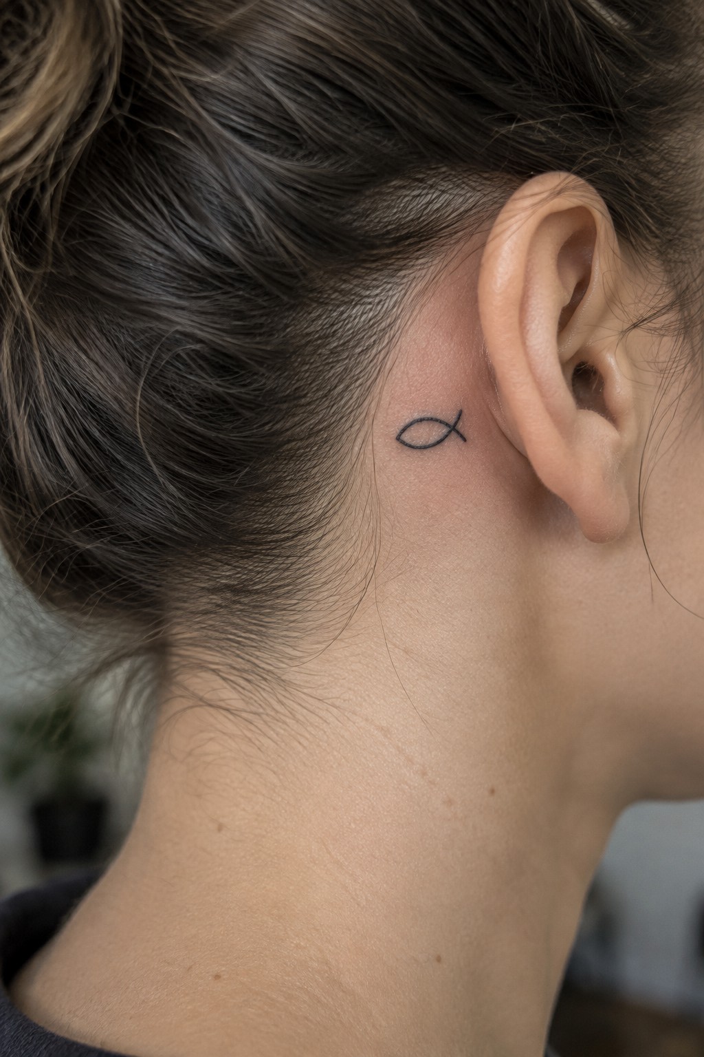 Small black outline of a fish tattoo placed behind the ear near the hairline on a person's head.