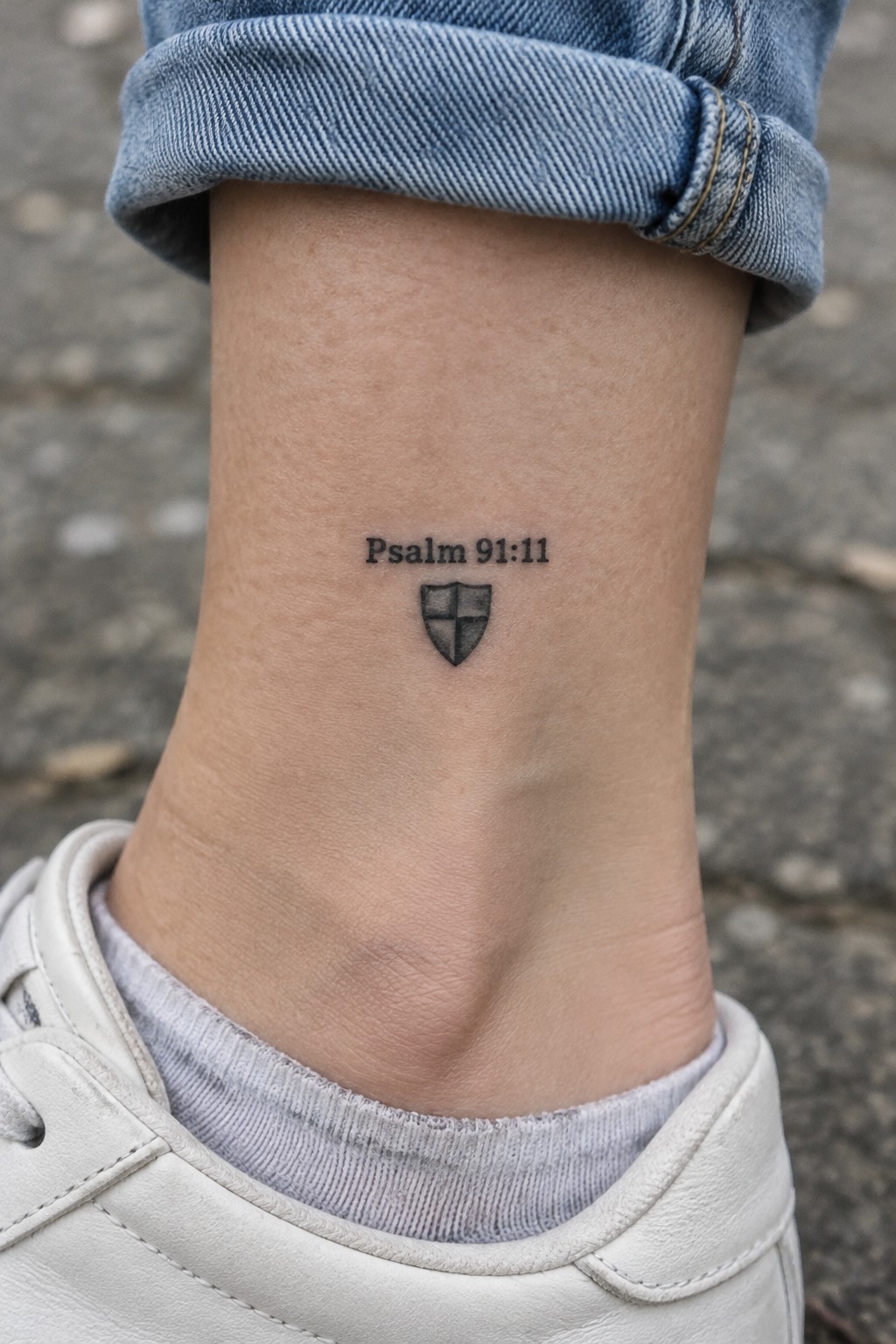 Psalm 91:11 tattoo above shield on ankle, rolled blue jeans, white sneaker.