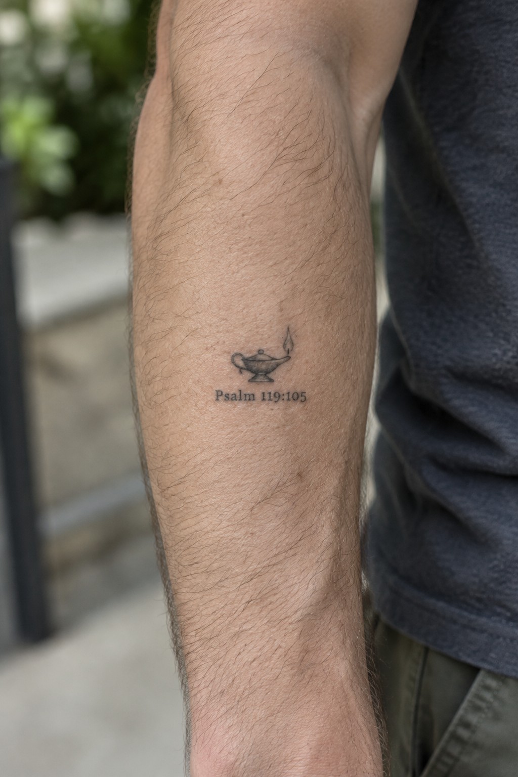 Small black ink tattoo of an oil lamp with flame and the text Psalm 119:105 on a person's forearm