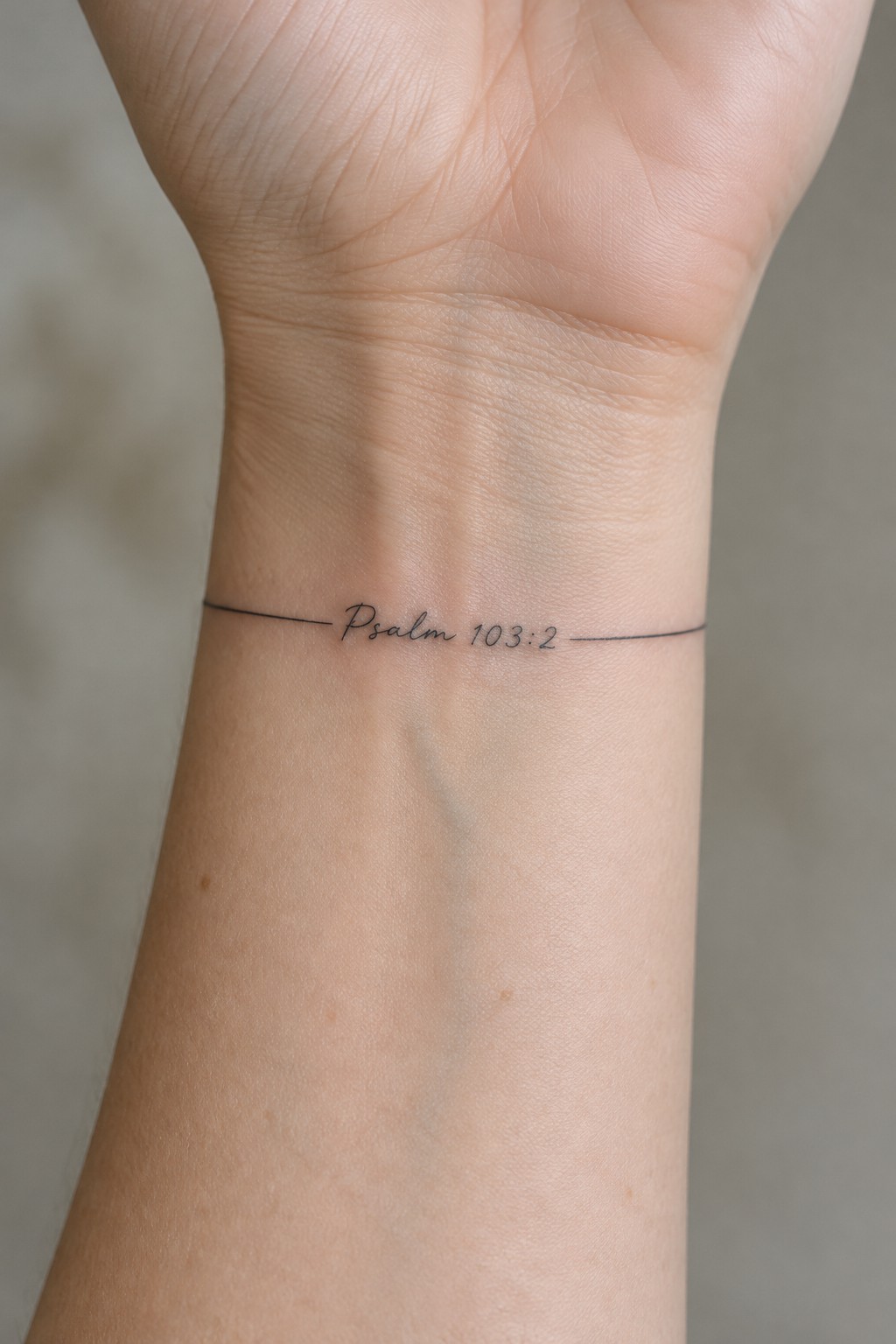 Wrist with fine-line black tattoo reading Psalm 103:2 between thin lines