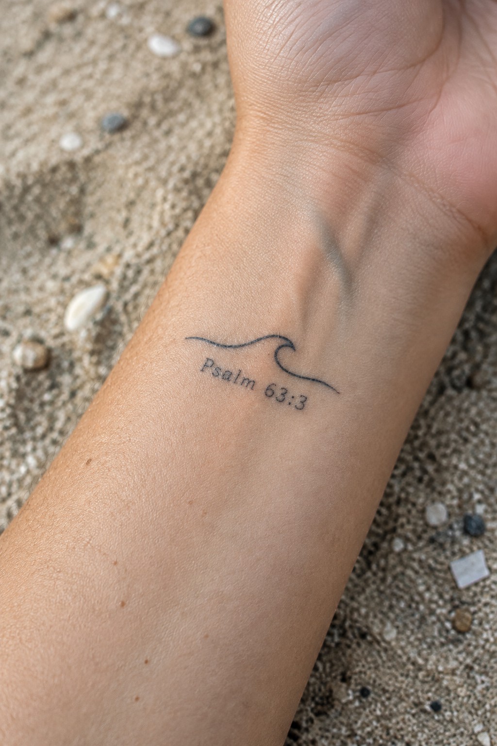 Wave tattoo with Psalm 63:3 text on wrist over beach sand