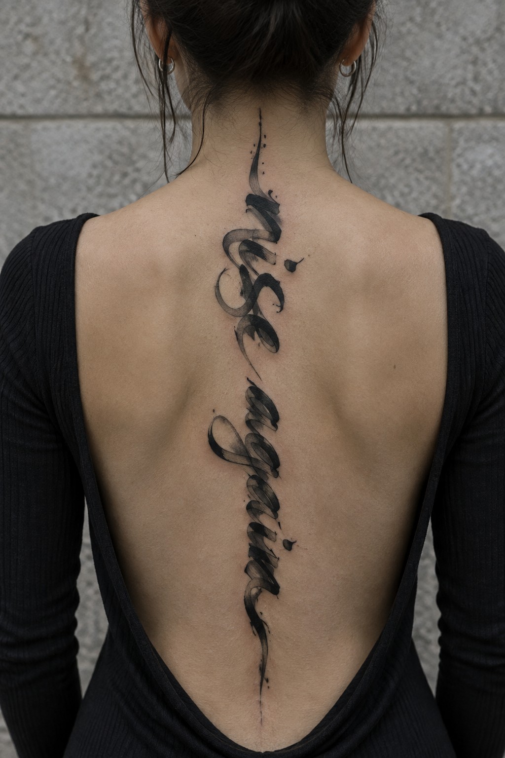 Woman with a long black calligraphy quote tattoo running down the center of her back from the nape toward the lower spine.