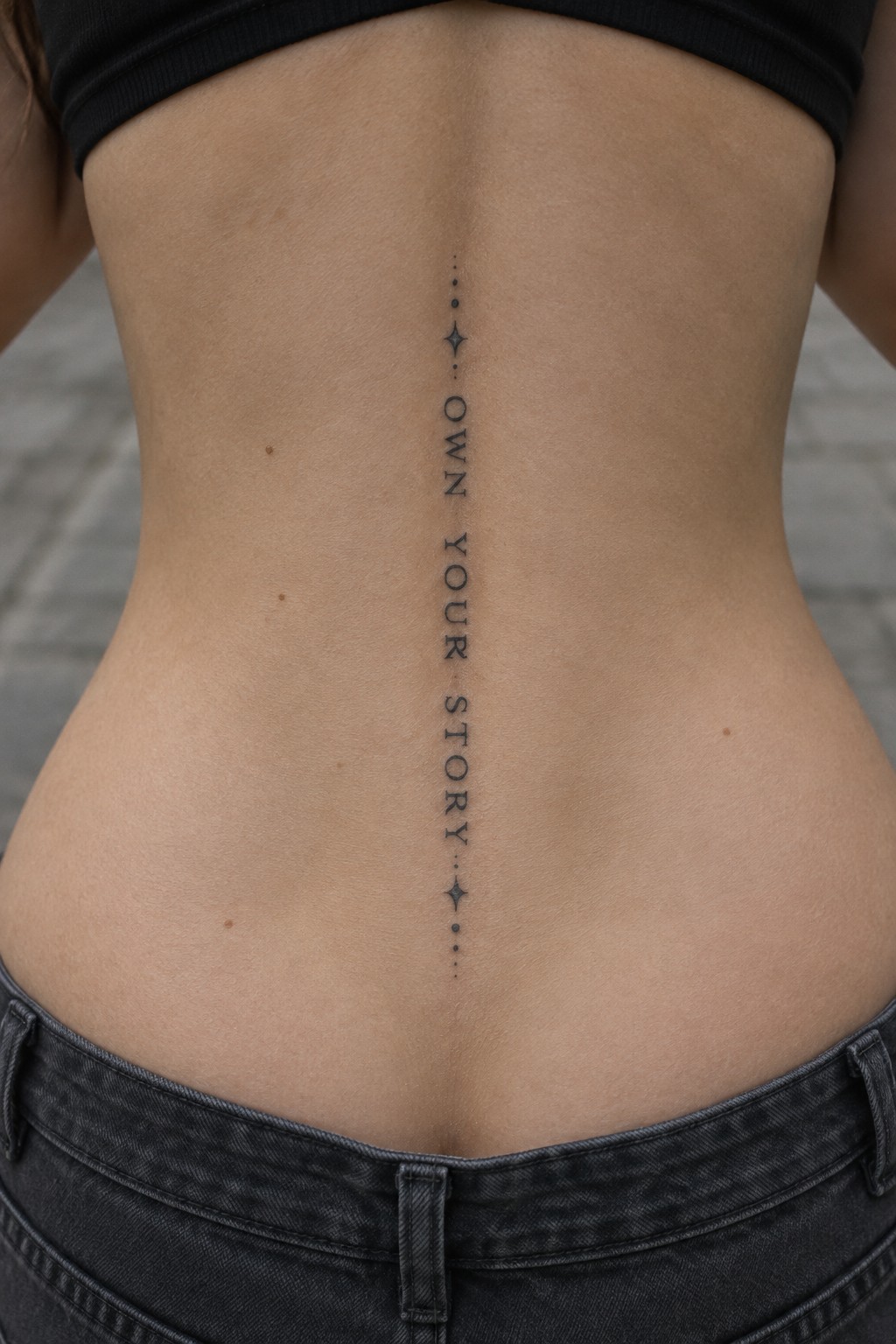 Vertical “Own Your Story” tattoo in fine black lettering down the center of a woman’s spine, with small star and dot details.