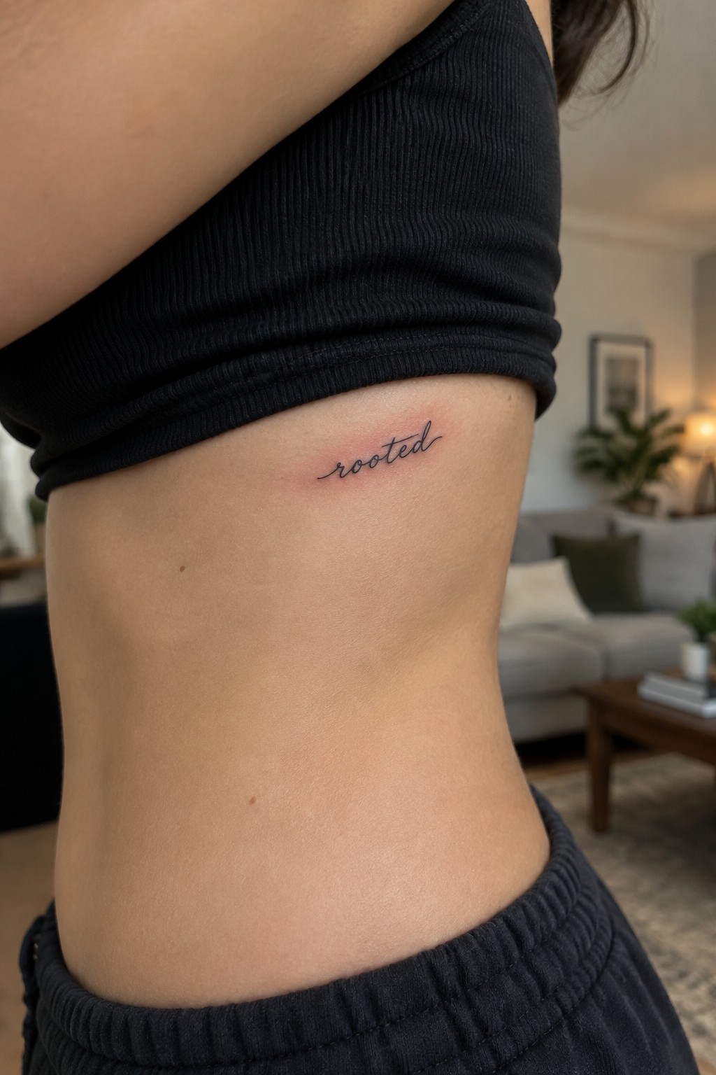 Small black cursive “rooted” tattoo on the side of a woman’s torso below a black top.