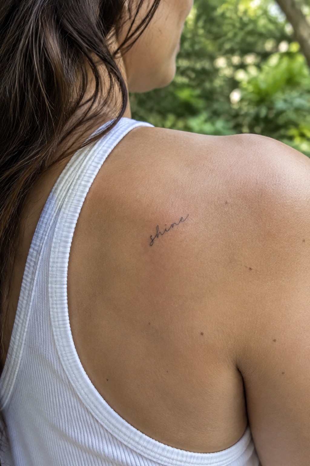 Small fine-line script tattoo reading “shine” on the upper shoulder above a white tank top.