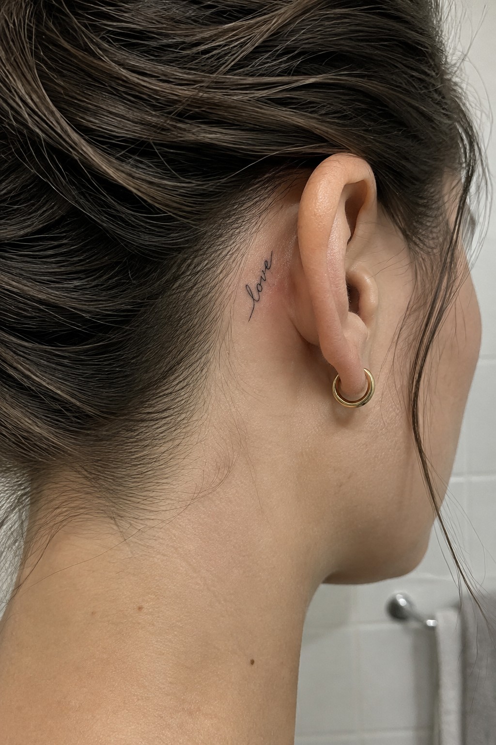 Small black script tattoo reading “love” behind a woman’s ear, above a gold hoop earring.