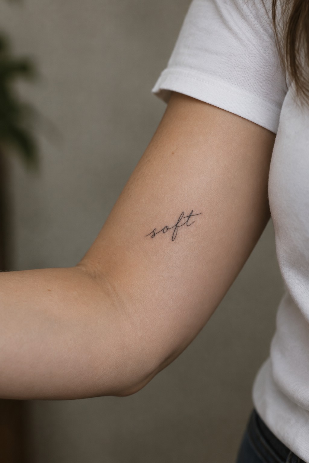 Small black script tattoo reading “soft” on the inner upper arm of a person wearing a white shirt.