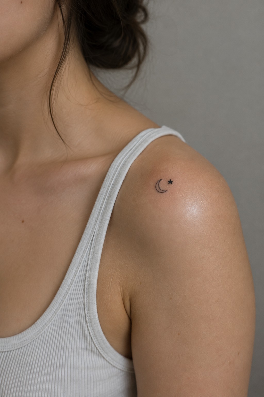 Small black crescent moon and star tattoo on a woman’s upper shoulder above a white tank top.