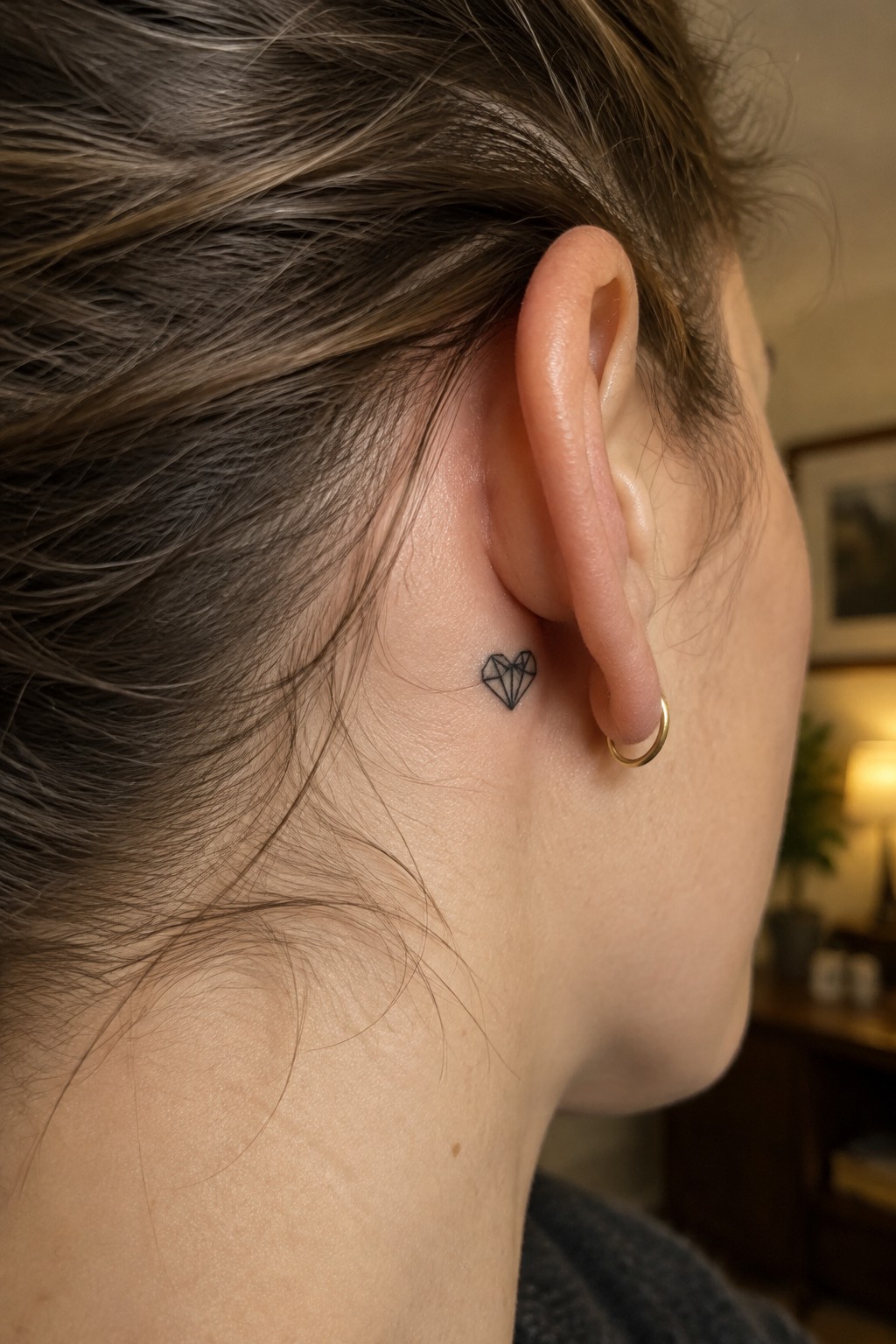 Small black geometric heart-shaped diamond tattoo behind a woman’s ear, beside a gold hoop earring.