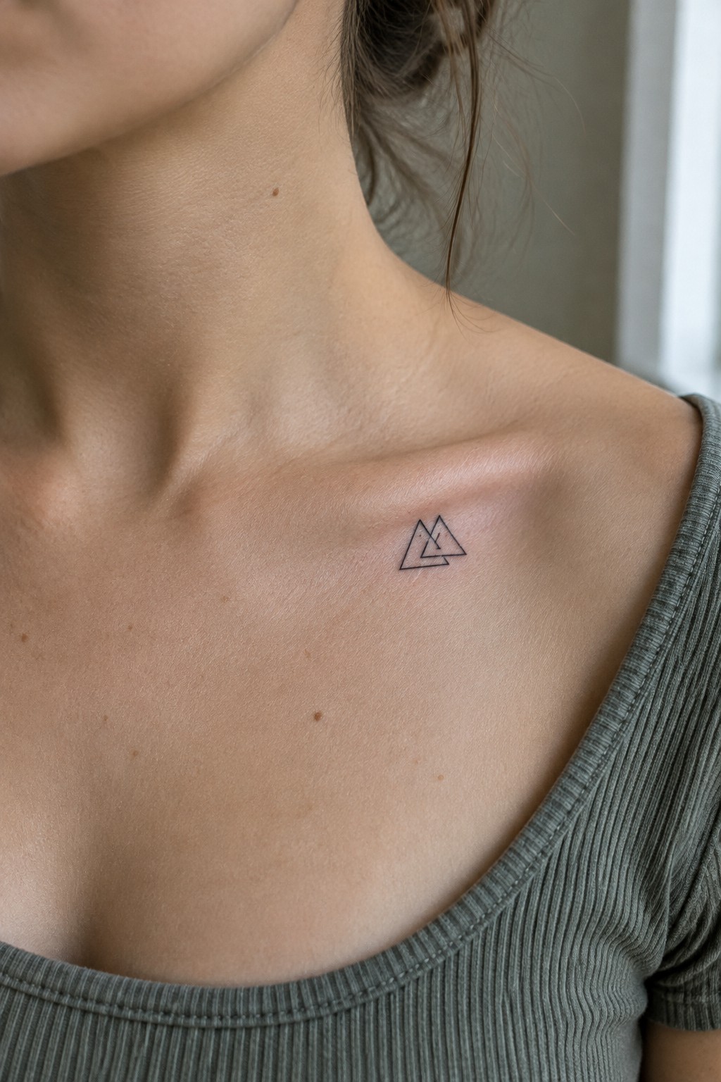 Small black outline tattoo of overlapping triangles on a woman’s upper chest near the collarbone.