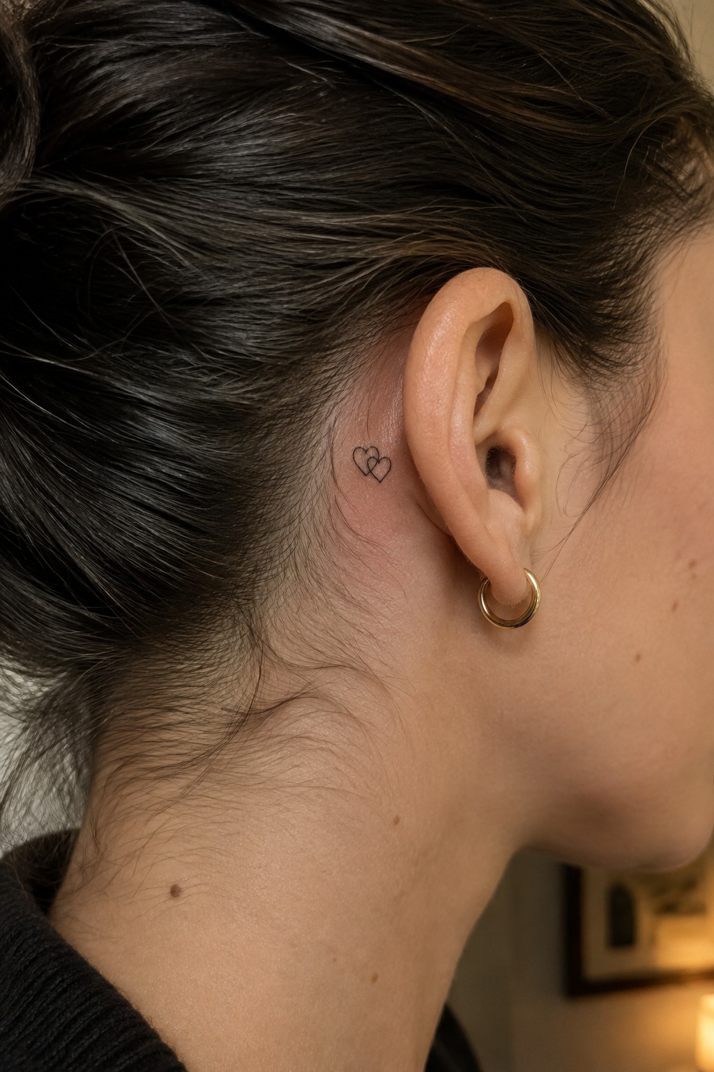 Small black outline tattoo of two overlapping hearts behind a woman’s ear.