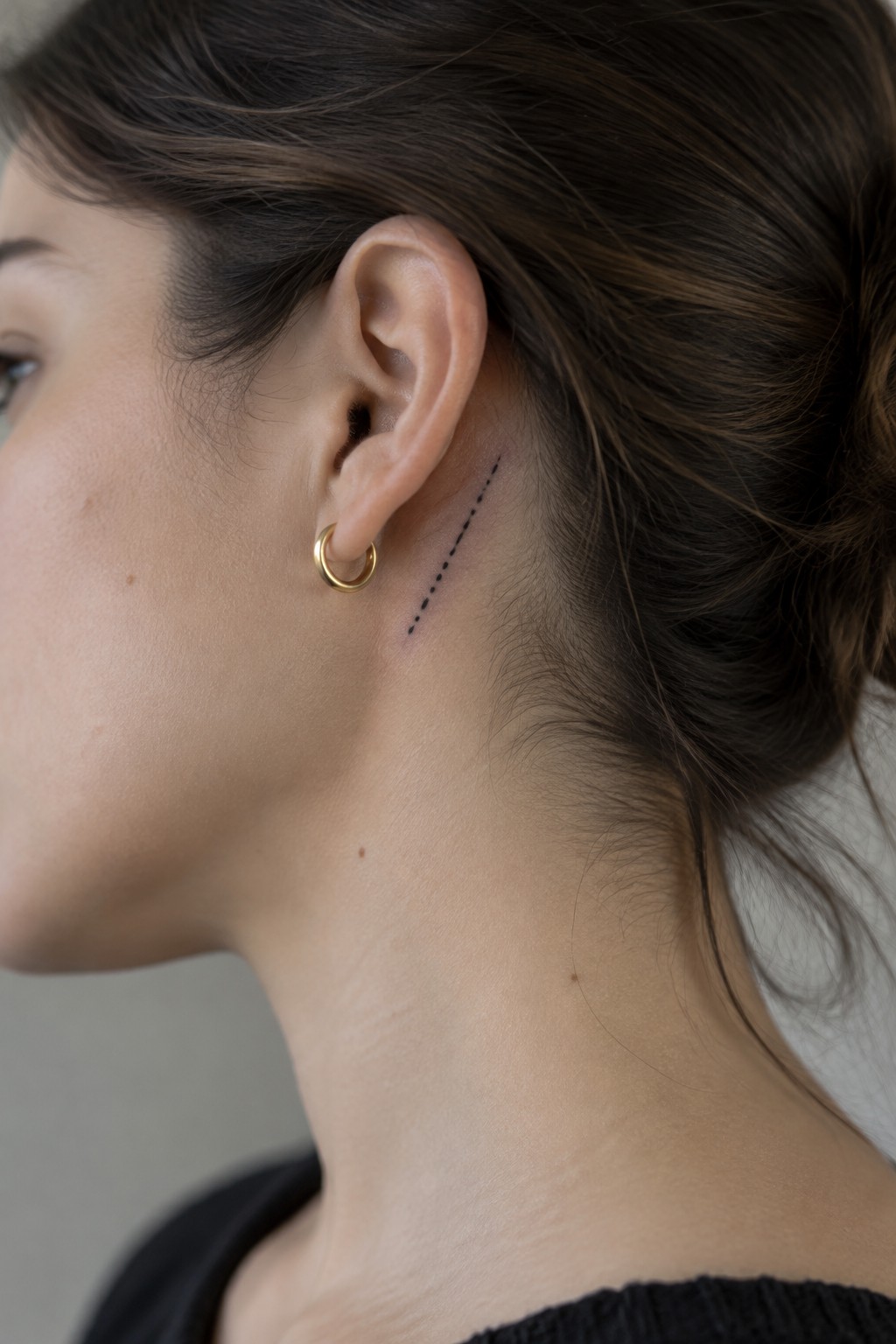 Close-up of a woman’s ear with a small black dotted line tattoo angled behind it and a gold hoop earring.
