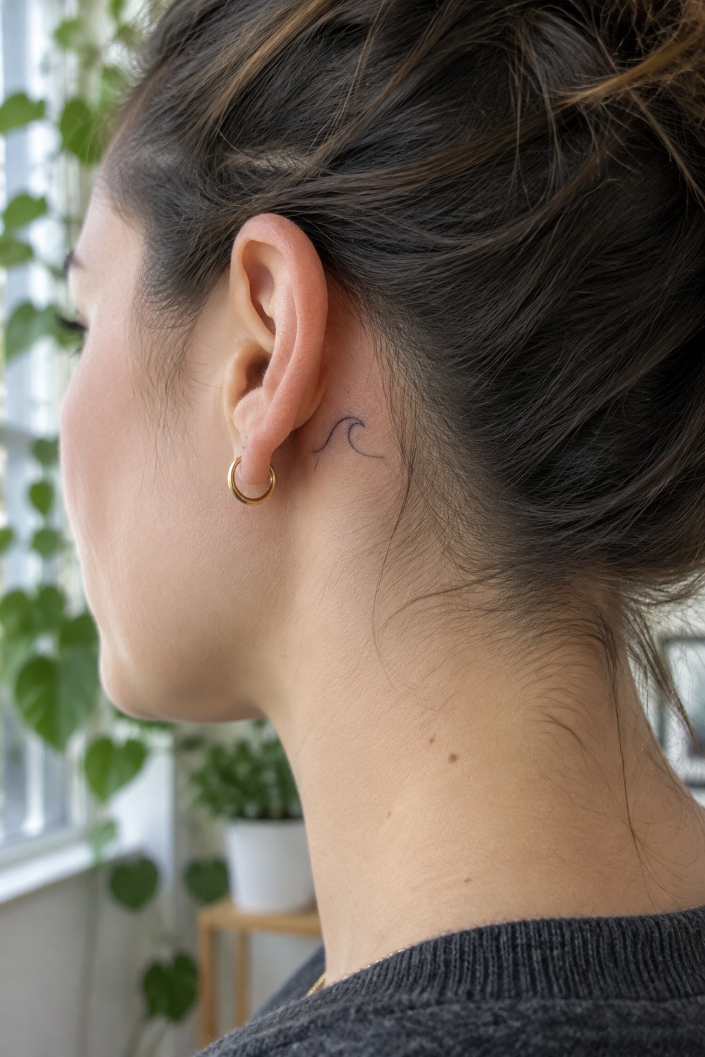 Small fine-line wave tattoo behind a woman’s ear, above a gold hoop earring.