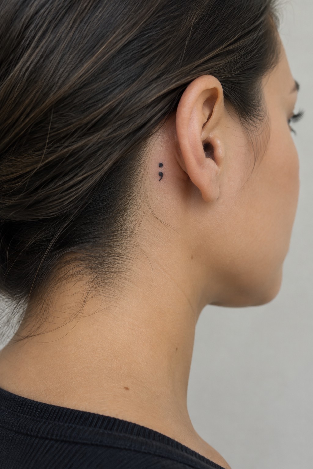 Small black semicolon tattoo behind a woman’s ear, set against her dark hair.