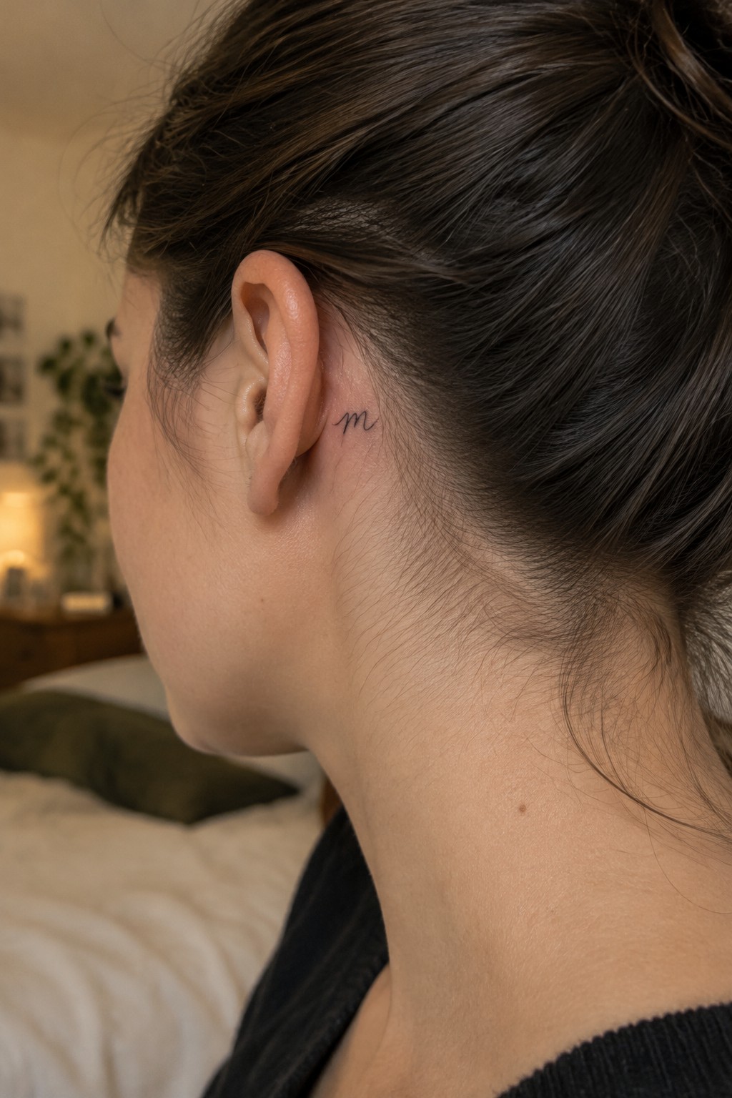 Small black lowercase m tattoo in fine script behind a woman’s ear.