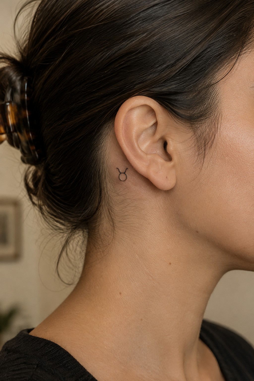 Small black outline Taurus symbol tattoo behind a woman’s ear.