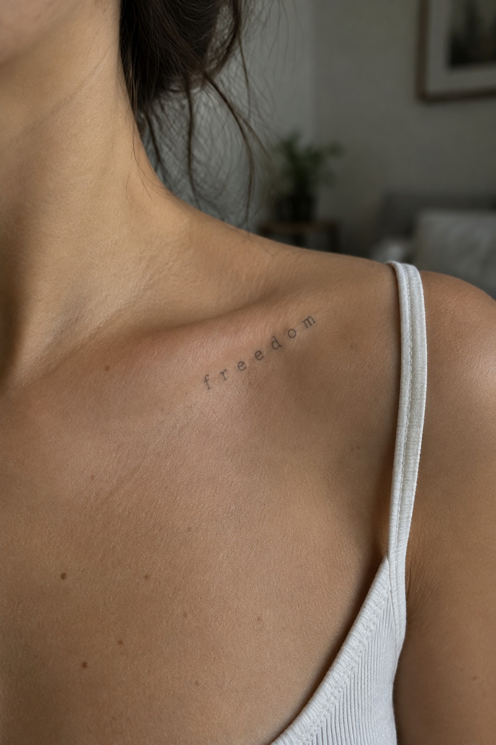 Small lowercase “freedom” tattoo in fine lettering along a woman’s collarbone, above a white tank top strap.