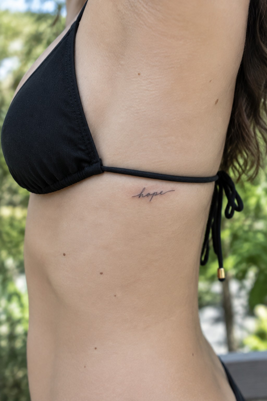 Small cursive “hope” tattoo on the side of a woman’s rib cage beneath a black bikini strap.