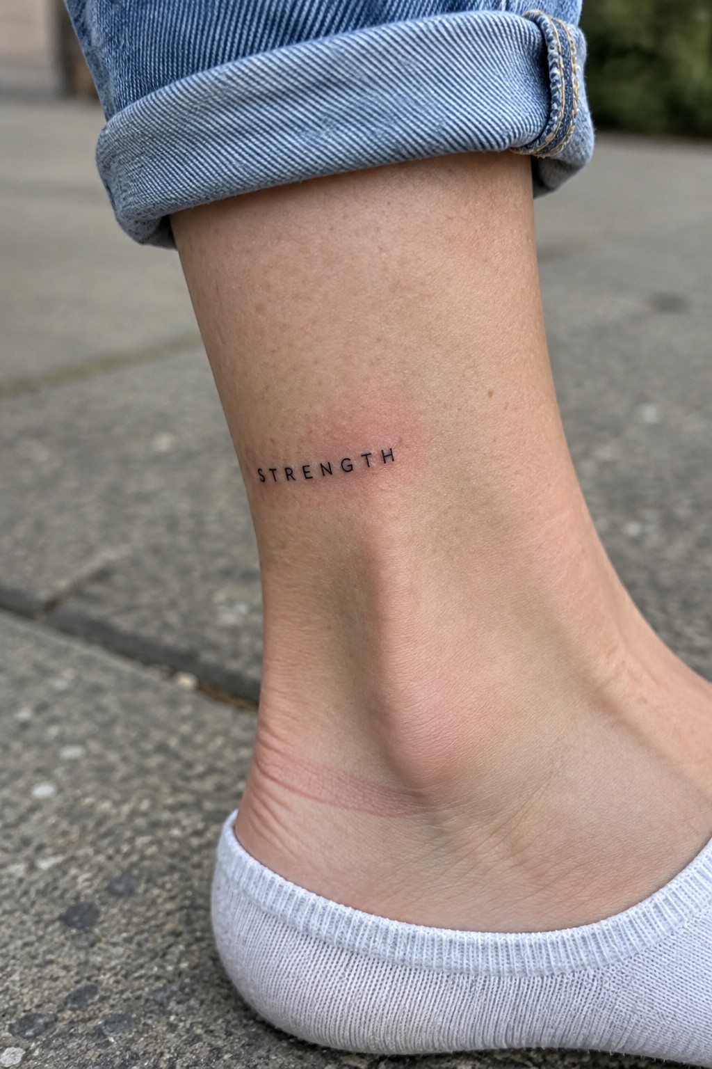 Small black uppercase “STRENGTH” tattoo on the inner ankle above a white sock.