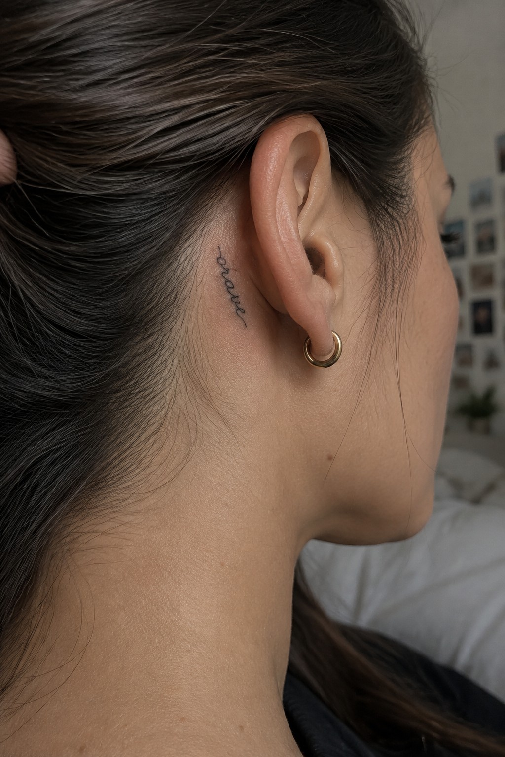 Small fine-script “brave” tattoo behind a woman’s ear, above a gold hoop earring.