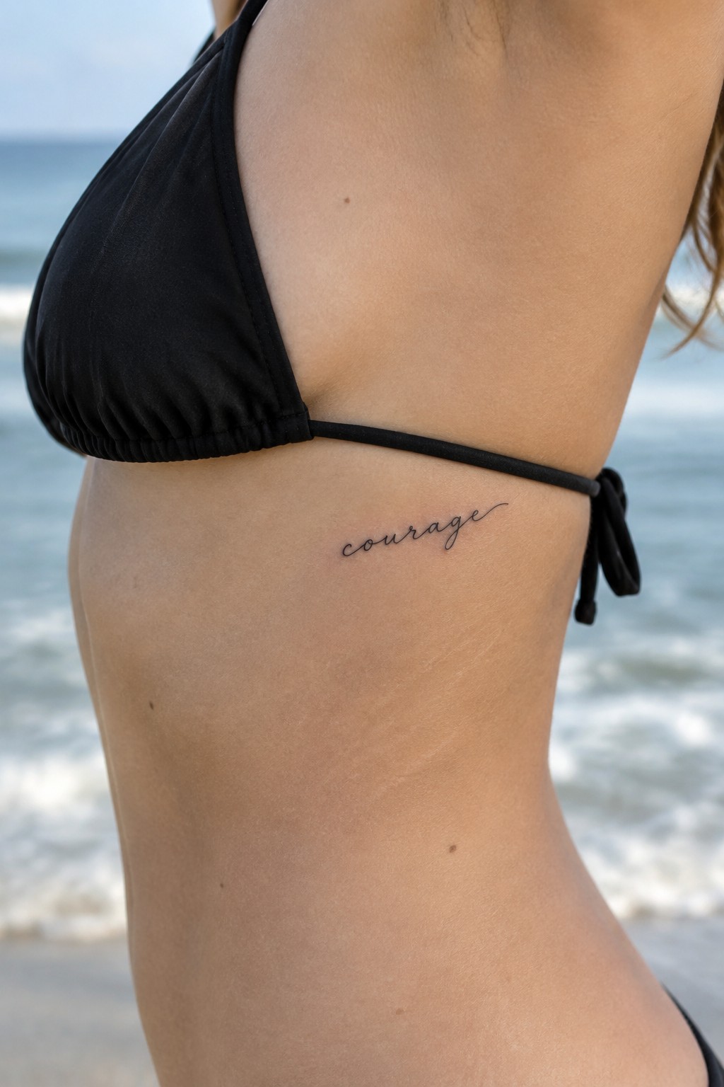 Small black cursive “courage” tattoo on the side of a woman’s torso below a black bikini top at the beach.