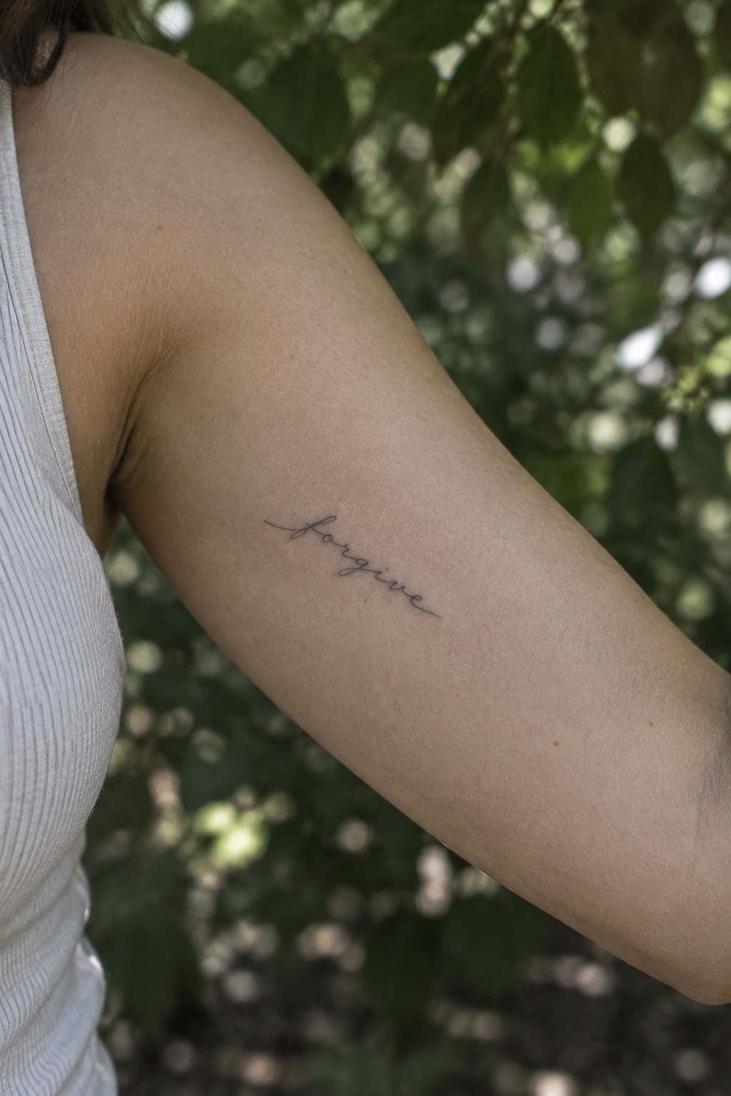 Small fine-line cursive “forgive” tattoo angled across a woman’s upper arm.