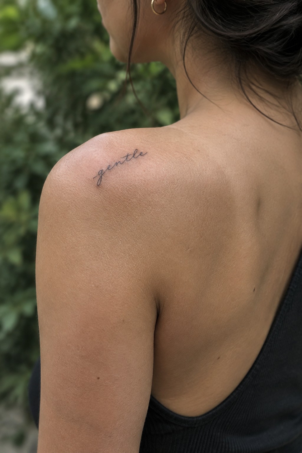 Small fine-line “gentle” script tattoo on a woman’s upper shoulder.