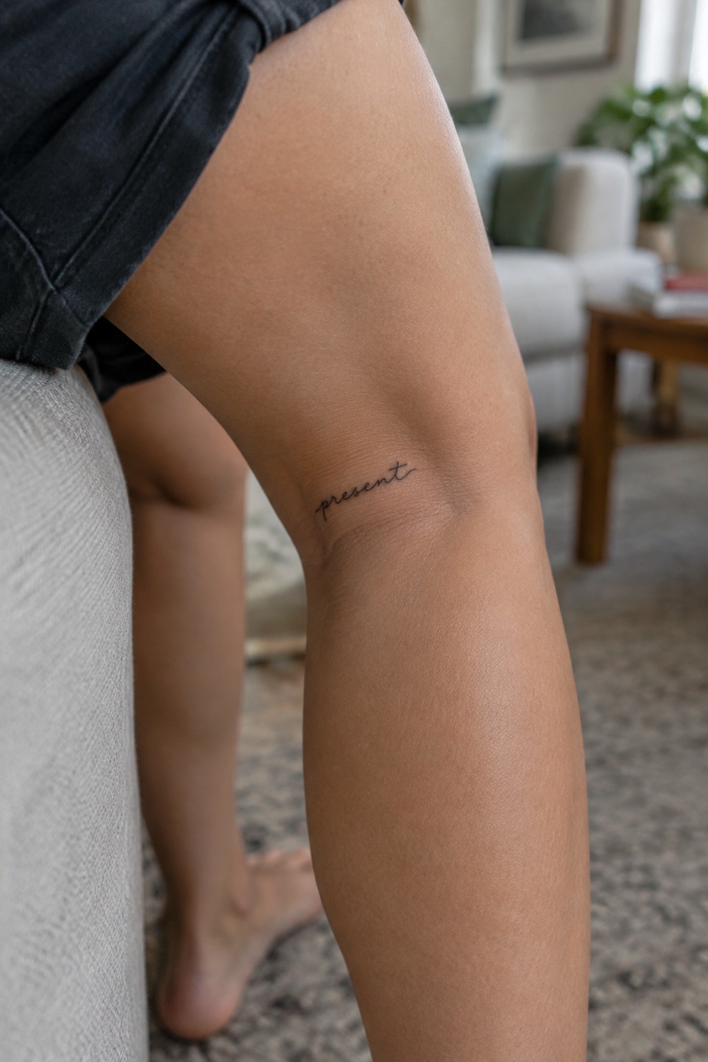 Small black cursive “present” tattoo above the back of the knee.