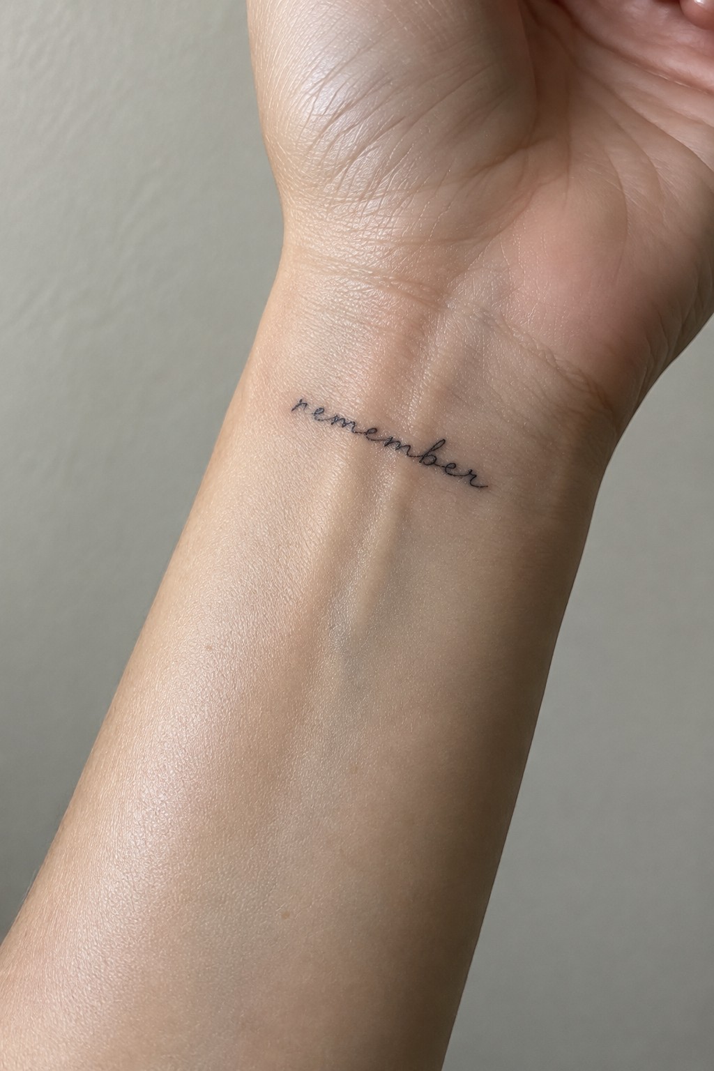 Small black cursive “remember” tattoo on the inner wrist.