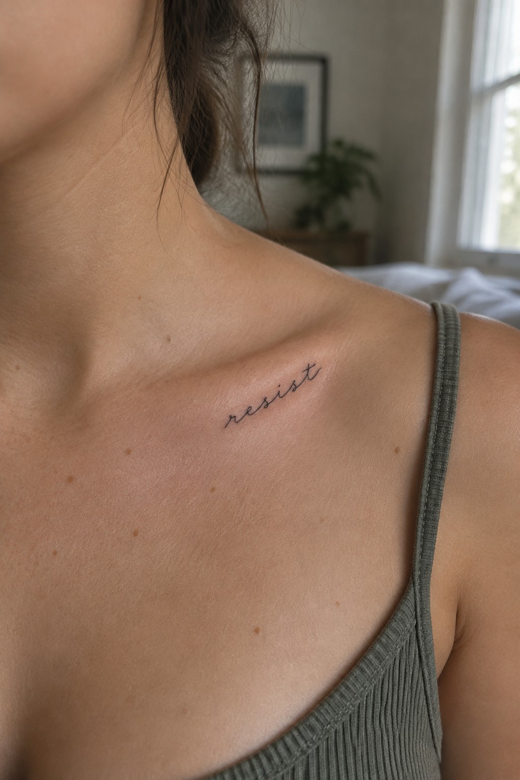 Small black “resist” script tattoo angled along a woman’s collarbone.