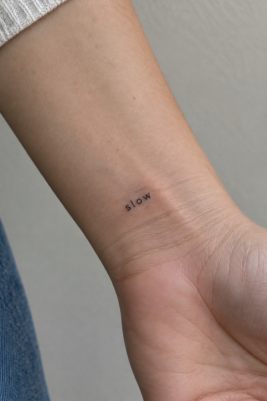Small black “slow” word tattoo on the inner wrist in spaced lettering.
